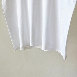 May include: A close-up of a white t-shirt, showing the fabric's texture and folds. The shirt is plain and appears to be made of a soft material. The bottom hem is visible, with a clean, simple finish. The background is a light, neutral color.