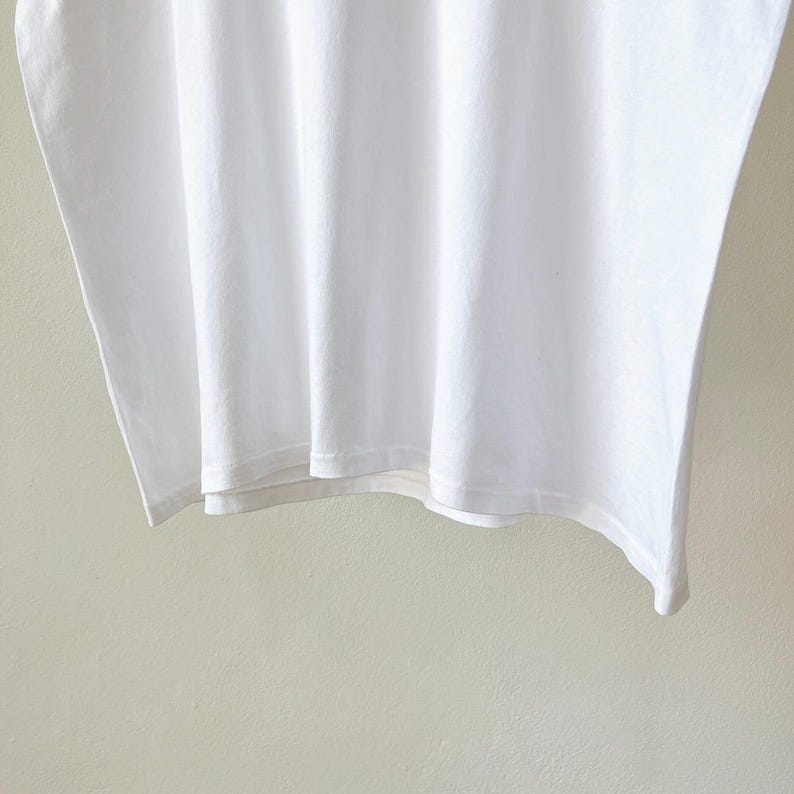 May include: A close-up of a white t-shirt, showing the fabric's texture and folds. The shirt is plain and appears to be made of a soft material. The bottom hem is visible, with a clean, simple finish. The background is a light, neutral color.