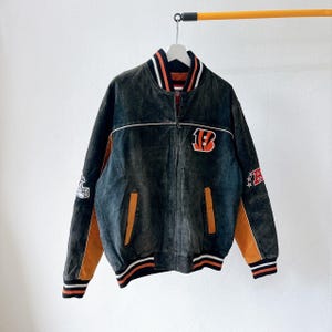 May include: A black and orange zip-up jacket featuring a large orange and black B logo on the chest, striped ribbed cuffs and collar, and embroidered patches on the sleeves. The jacket has two front pockets and a full-length front zipper closure.