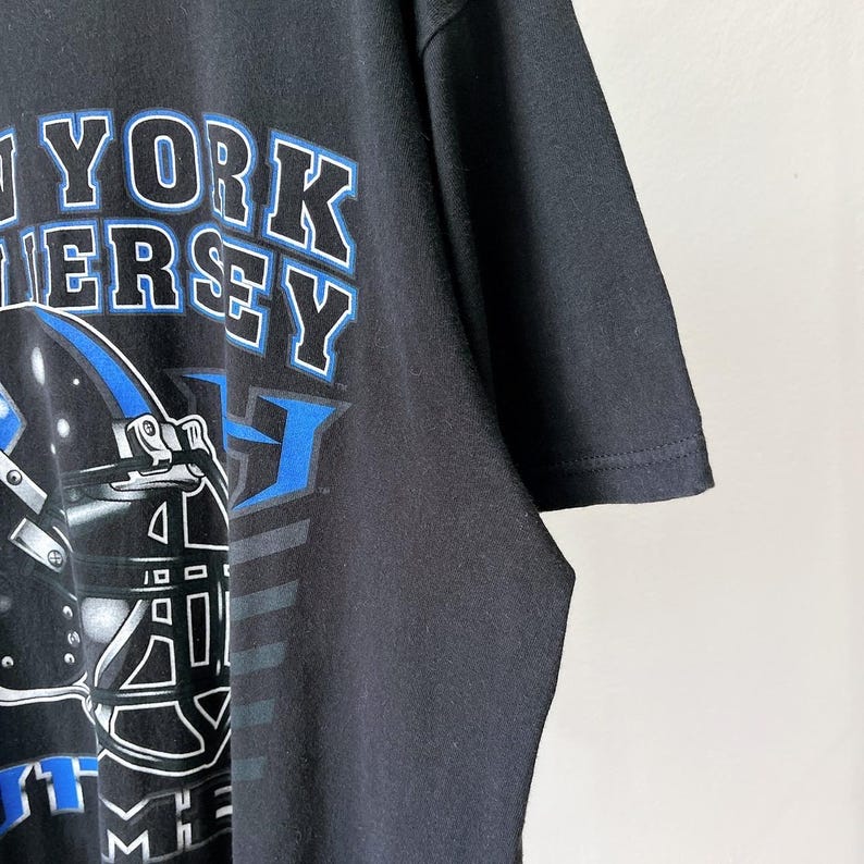 Puede incluir: Camiseta negra con un dise&ntilde;o gr&aacute;fico con las palabras "NEW YORK JERSEY" en letras blancas y azules. El dise&ntilde;o incluye un casco de f&uacute;tbol con detalles en azul. La camiseta est&aacute; hecha de un material suave.