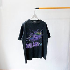 Może przedstawiać: Czarny t-shirt z graficznym wzorem z fioletowym napisem "DEVIL RAYS" i stylizowanym wizerunkiem płaszczki. Projekt zawiera wybuch fioletowych i zielonych kolorów. Koszulka wisi na białym wieszaku.
