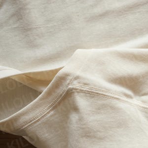 May include: Close-up of a cream-colored sweatshirt. The image highlights the neckline and shoulder seam, showcasing the fabric's texture and stitching details. The garment appears to be made of a soft, comfortable material.