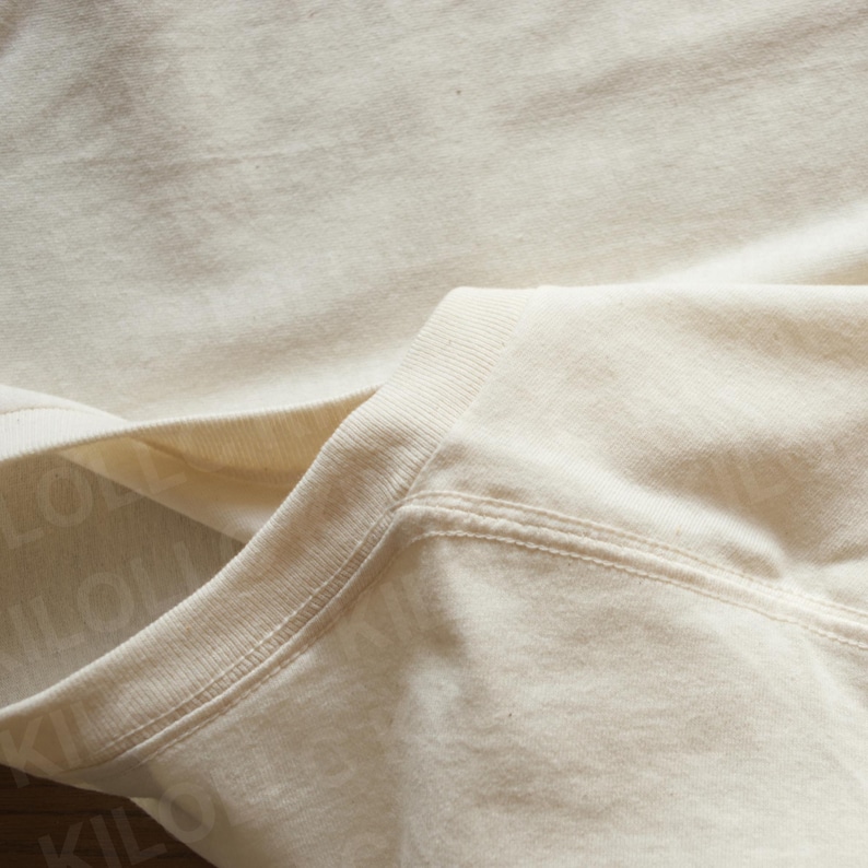 May include: Close-up of a cream-colored sweatshirt. The image highlights the neckline and shoulder seam, showcasing the fabric's texture and stitching details. The garment appears to be made of a soft, comfortable material.