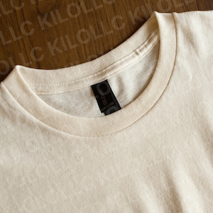 May include: Close-up of a cream-colored t-shirt with a crew neck. The shirt has a black tag with the brand name "GILDAN" printed on it. The fabric appears to be a soft, medium-weight cotton.