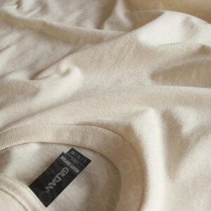 May include: Close-up of a cream-colored t-shirt with a crew neck. The fabric is slightly wrinkled, and a black tag with the brand name "GILDAN" and "HEAVY COTTON" is visible.