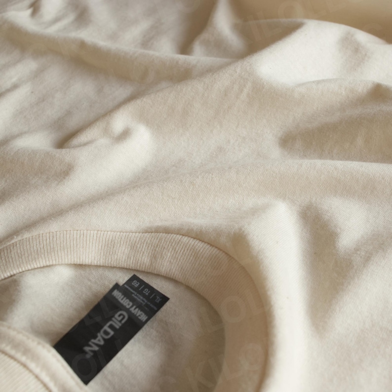 May include: Close-up of a cream-colored t-shirt with a crew neck. The fabric is slightly wrinkled, and a black tag with the brand name "GILDAN" and "HEAVY COTTON" is visible.
