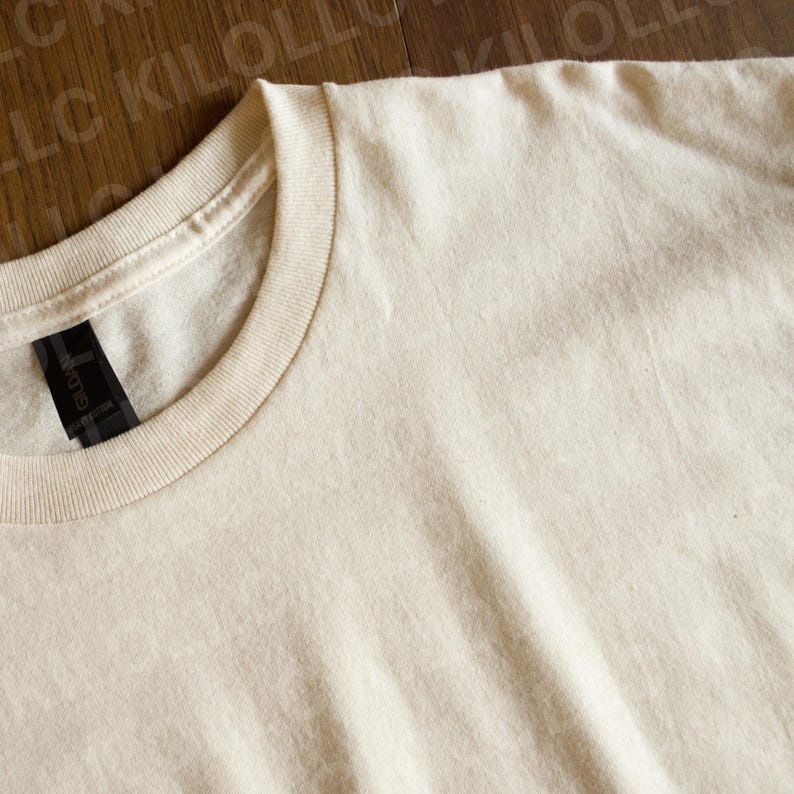 May include: Close-up of a cream-colored t-shirt. The shirt has a crew neck and short sleeves. The tag on the inside of the collar is black with white text. The fabric appears to be soft and slightly textured.