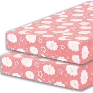 May include: Two pink fitted cot sheets featuring a repeating pattern of white smiling clouds, stars, and crescent moons. The sheets are made of a soft material, designed for a baby's cot. The overall theme is a whimsical, nighttime sky.