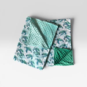 May include: A square baby blanket featuring a white fabric top with a repeating pattern of green and blue sea turtles. The blanket has a textured, dimpled minky backing in a light mint green colour, with a corner folded over to reveal the texture.