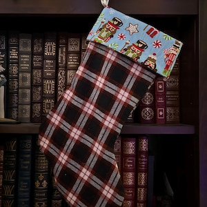 Peut inclure: Une chaussette de Noël avec un motif à carreaux noir, rouge et blanc. Le revers est en tissu bleu clair avec des illustrations de casse-noisettes, d'étoiles et de cannes en sucre. Un ruban blanc est attaché pour l'accrocher.