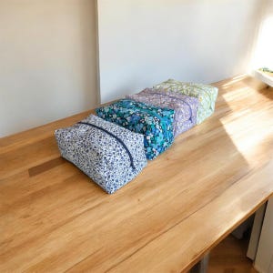 Peut inclure: Quatre trousses rectangulaires en tissu matelassé disposées en rangée. Les trousses présentent divers motifs floraux dans des tons de bleu, violet et vert, chacune équipée d'une fermeture éclair assortie sur le bord supérieur.