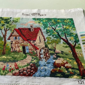 May include: Needlepoint of a quaint cottage scene with a red roof, water wheel, and flowing stream. The artwork is on white canvas, with the text "ROYAL PARIS" at the top. The color palette includes greens, blues, and browns.