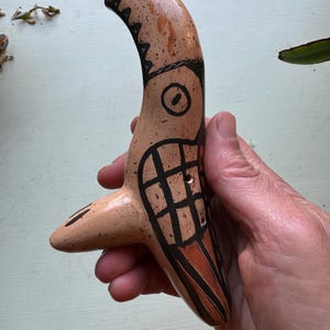 Può includere: Un ocarina in ceramica dipinta a mano a forma di uccello con disegni neri e marroni su uno sfondo marrone chiaro.