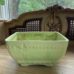 Pode incluir: Um vaso retangular de cerâmica verde claro com borda ondulada e design de folhas. O vaso tem uma aparência ligeiramente desgastada, sugerindo idade ou estilo vintage. Adequado para uso interno ou externo.