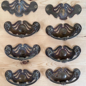 Puede incluir: Un conjunto de tiradores y cubiertas de cerradura antiguos de bronce. Los tiradores tienen un diseño de asa curvada con detalles decorativos. Las cubiertas de cerradura tienen un diseño ornamental y simétrico. La ferretería está envejecida.