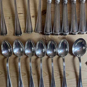 May include: A collection of antique silverware, including spoons, knives, and forks, displayed on a wooden surface. The silverware has a polished, silver appearance, reflecting light. The spoons have rounded bowls, and the knives have decorative handles.