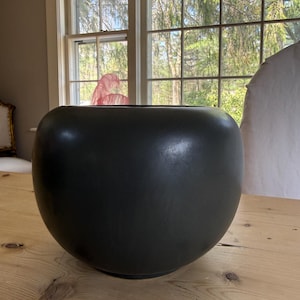 Può includere: Una grande ciotola in ceramica nera opaca dalla forma arrotondata. La ciotola è appoggiata su un tavolo di legno chiaro, con una finestra e alberi visibili sullo sfondo. La superficie liscia della ciotola contrasta con le venature naturali del legno.