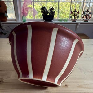 May include: A large, reddish-brown ceramic bowl with vertical cream-colored stripes. The bowl has a wide, rounded shape and sits on a light-colored wooden surface. The stripes are evenly spaced and run from the rim to the base.