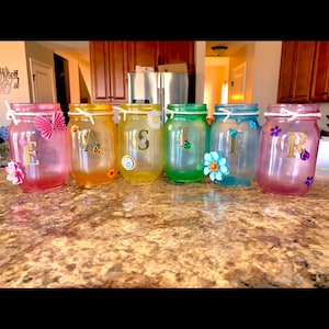 May include: Six decorative mason jars in various colors, each adorned with a letter and floral or butterfly embellishments. The jars are arranged on a countertop, showcasing a personalized theme.