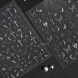 May include: Two sheets of silver nail art stickers featuring crosses, butterflies, moons, hearts, and stars. The stickers are on a clear backing, set against a dark background. The metallic stickers have a 3D effect, ideal for nail art.