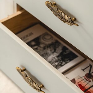 Puede incluir: Primer plano de un armario gris claro con un caj&oacute;n abierto, que revela un libro en su interior. El armario tiene dos tiradores de caj&oacute;n en forma de pluma de color bronce. El caj&oacute;n abierto revela un libro con la palabra "GOGH" visible.