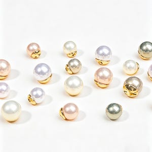 May include: An assortment of faux pearl beads in various colors, including white, pink, and gray. Each bead has a gold-colored base. The beads are scattered on a white surface, creating a visually appealing arrangement.