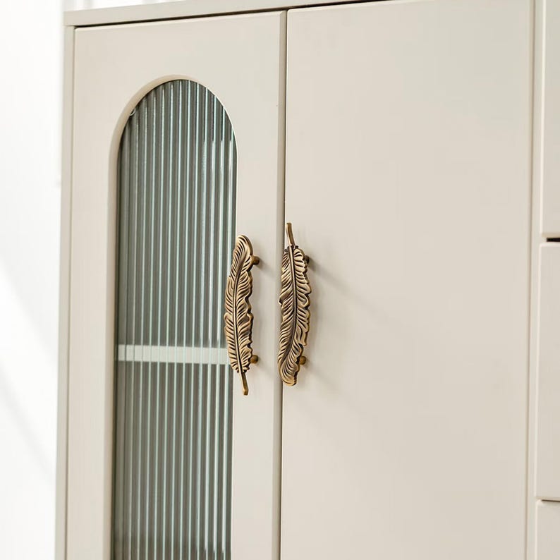 Puede incluir: Puertas de armario color crema con tiradores de bronce en forma de pluma. Una puerta presenta un panel de vidrio con detalles acanalados verticales. El armario forma parte de un mueble m&aacute;s grande.
