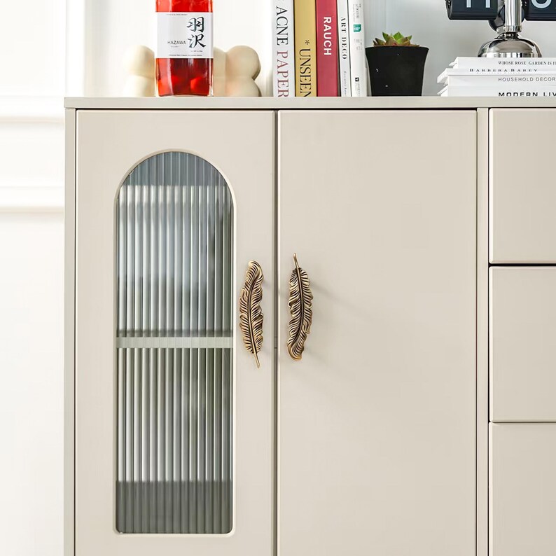 Puede incluir: Armario color crema con un panel de puerta de cristal y dos tiradores dorados en forma de pluma. El panel de cristal tiene l&iacute;neas verticales. El armario forma parte de un mueble m&aacute;s grande, con libros y decoraci&oacute;n encima.