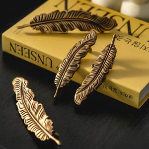 May include: Close-up of several bronze-colored feather-shaped cabinet pulls and drawer handles. The hardware is arranged on and around a yellow book. The feather design has detailed veining and a vintage aesthetic.
