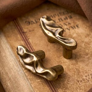 May include: Two cabinet knobs with an irregular, organic, and flowing shape. The knobs feature a metallic finish with a dark, antique brass-like patina, showcasing deep grooves and raised ridges that create a textured, sculptural appearance.