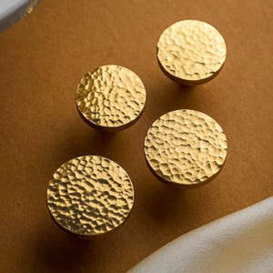 May include: A set of four circular cabinet knobs with a metallic gold finish. Each knob features a distinct hammered texture across the flat, round surface, creating a reflective and uneven appearance that catches the light from multiple angles.