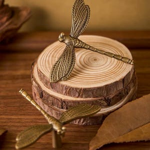 May include: Two antique brass-toned metal dragonfly drawer knobs featuring detailed wing textures and segmented bodies. One knob is positioned on top of a stack of three circular wood slices, while the second knob is placed in the foreground.