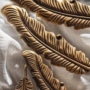 Puede incluir: Primer plano de varias piezas de herrajes decorativos en forma de pluma de color bronce. Las plumas tienen vetas detalladas y est&aacute;n dispuestas sobre una superficie blanca y texturizada. Los herrajes parecen ser de metal y tienen una est&eacute;tica vintage.