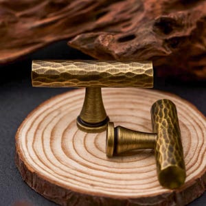 May include: Two antique brass cabinet pulls with a hammered texture. The T-shaped pulls have cylindrical handles and conical bases. They are displayed on a wooden slice, highlighting their vintage aesthetic.