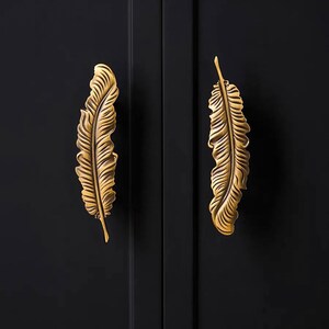 Puede incluir: Dos tiradores decorativos para armarios con forma de pluma. Los tiradores son de metal dorado y est&aacute;n montados en una puerta de armario negra. El dise&ntilde;o de la pluma tiene vetas detalladas y una superficie texturizada.