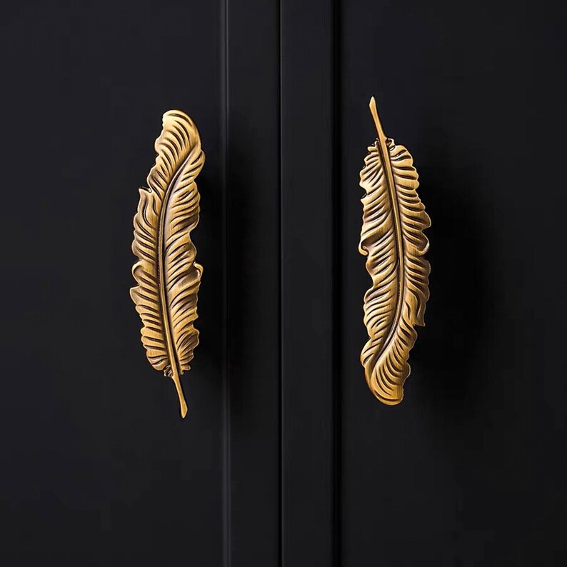 Puede incluir: Dos tiradores decorativos para armarios con forma de pluma. Los tiradores son de metal dorado y est&aacute;n montados en una puerta de armario negra. El dise&ntilde;o de la pluma tiene vetas detalladas y una superficie texturizada.