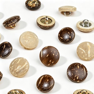 Può includere: Una collezione di bottoni rotondi in varie tonalità di marrone e beige, sparsi su una superficie bianca. Alcuni bottoni hanno un occhiello metallico, altri un gambo. I bottoni sembrano essere fatti di materiali naturali, forse cocco, con un diametro di circa 2 cm.