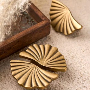 May include: Two brass-colored metal cabinet pulls shaped like open fans. Each pull features a semi-circular design with radiating embossed lines that extend from the center to the scalloped outer edge, creating a textured, metallic finish.
