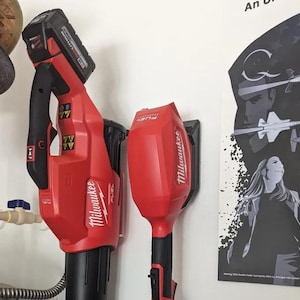 Puede incluir: Dos herramientas eléctricas inalámbricas Milwaukee FUEL rojas y negras montadas en una pared blanca. Las herramientas incluyen un soplador de hojas y un soplador de mano. Una batería negra y roja está conectada al soplador de hojas. Un póster con un gráfico en blanco y negro es visible en el fondo.