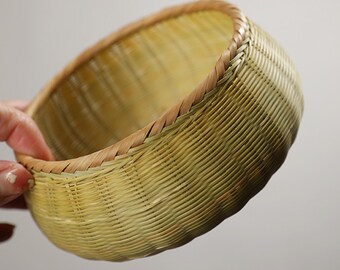 May include: A hand-woven, round basket made of light green and brown bamboo. The basket has a woven design with a brown rim. The basket is held by a hand, and the background is a neutral color.