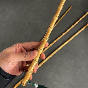 May include: A light brown bamboo wand with decorative joints and a green tassel. The wand is held in a hand, with several other similar wands in the background. The wands have a natural, textured appearance.