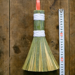 May include: A small, handcrafted broom with a green and yellow straw head and a bamboo handle. The handle is wrapped with white cord and has a red ribbon loop for hanging. The broom is approximately 25 cm long.