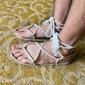 May include: Handmade sandals with woven straw soles and white rope straps. The rope crisscrosses over the foot and ties around the ankle. The sandals are designed for comfort and style, perfect for warm weather.