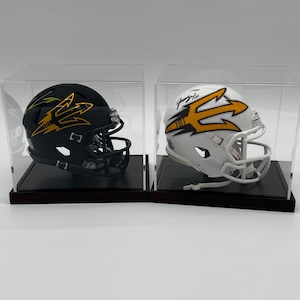May include: Two miniature football helmets, one black with gold accents and the other white with gold accents, are displayed in clear acrylic cases. Each helmet features a stylized trident logo. The helmets sit on dark wooden bases.