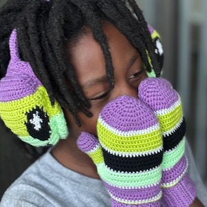May include: A person wearing a matching crochet hat and mittens set. The hat and mittens are purple, green, black, and white striped. The mittens are covering the person's face. The hat has a cartoon eye design.