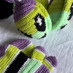 May include: Crocheted ear muffs and fingerless gloves in a vibrant color scheme. The ear muffs feature a purple and lime green design with black accents. The gloves have a striped pattern of purple, black, white, and lime green. Yarn balls are in the background.
