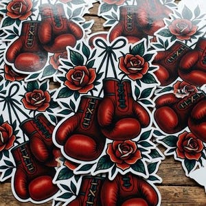May include: Red boxing glove stickers with a traditional tattoo design. Each glove is paired with red roses and green leaves. The stickers are on a wooden surface.