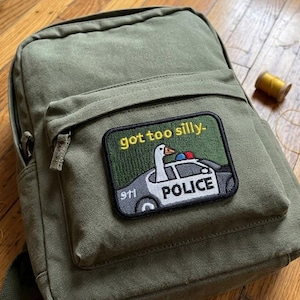 May include: Olive green canvas backpack with a patch that reads "got too silly-" featuring a cartoon goose in a police car. Includes a zipper closure, a small spool of thread, and small scissors.