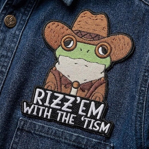 May include: Embroidered patch featuring a cartoon frog wearing a cowboy hat, jacket, and bow tie. The frog is green and white with brown accents. The patch includes the text "Rizz'em with the 'tism" in white letters on a black background. The patch is sewn onto a denim jacket.