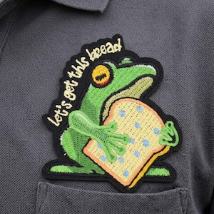 May include: A dark gray polo shirt with a patch featuring a green frog holding a slice of bread. The patch has the text "let's get this bread" in yellow. The frog has yellow eyes and is holding a slice of bread with blue dots.